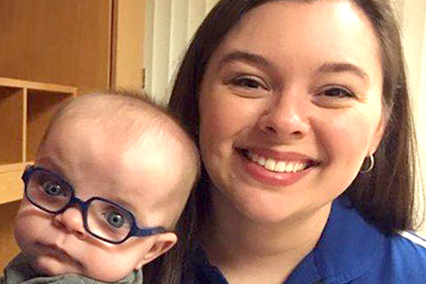 Close-up portrait of an infant wearing glasses beside an adult in an indoor clinical setting."