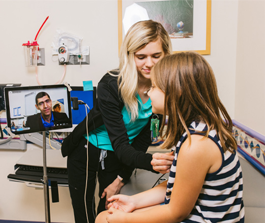 A woman uses a stethoscope to listen to the heart of a girl sitting on an exam room table. There is a tablet on a stand next to them and a man observes on the screen.