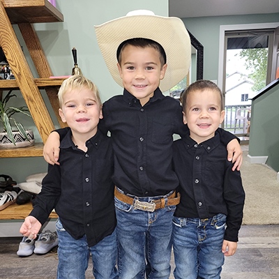 Three little boys in matching black shirts, blue jeans and brown shoes smile together. The oldest and tallest is in the middle with his arms around his younger brothers. 