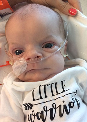Infant being held by a caregiver, wearing a nasal cannula for oxygen support and a shirt that reads 'Little Warrior.'