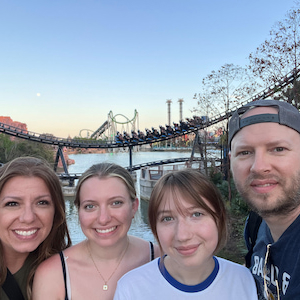 Four people posing for a selfie outdoors near a waterfront amusement park. A roller coaster and other rides are visible in the background, with water, a bridge, and a clear evening sky behind them.