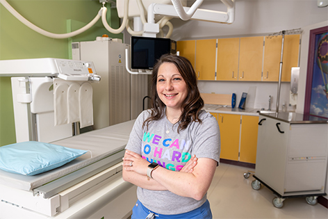 Person standing with arms crossed beside imaging equipment in a clinical exam room, with cabinets, medical equipment, and a monitor visible in the background.