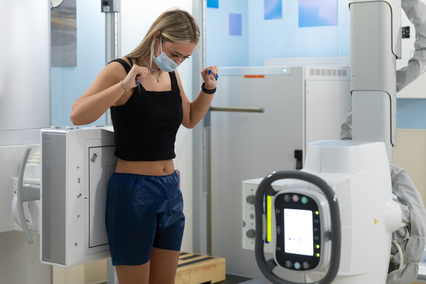 Patient standing in an imaging room preparing for an X-ray exam, with radiology equipment visible nearby.