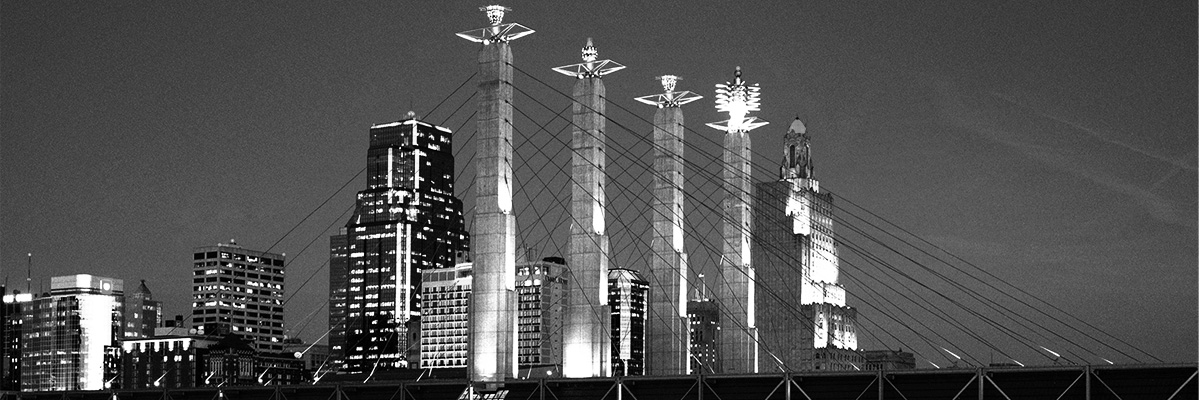 Alt text: Black-and-white photograph of the Kansas City skyline at night, featuring illuminated downtown buildings and the cable-stayed towers of the Christopher S. Bond Bridge in the foreground.