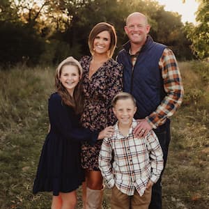 Family portrait of four people standing together outdoors in a grassy field during golden-hour light. Two adults stand behind two children, with trees and natural scenery in the background. The group is posed closely together for the photo.