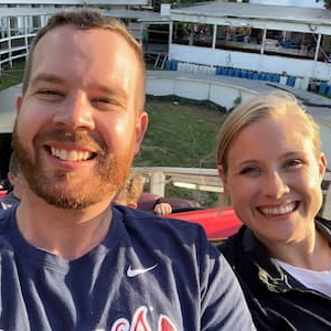 Close-up selfie of two people seated outdoors at a stadium or event venue. The photo shows both people smiling at the camera, with seating areas, railings, and part of the venue visible in the background.