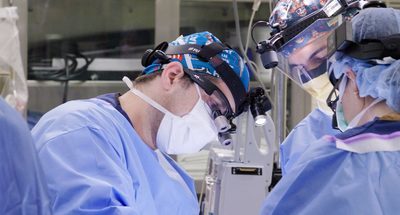 Medical team performing surgery in an operating room, with one lead surgeon focused on the procedure while others assist and observe. All are wearing surgical masks, gowns, and headgear with magnifying glasses. Medical equipment and monitors are visible in the background.
