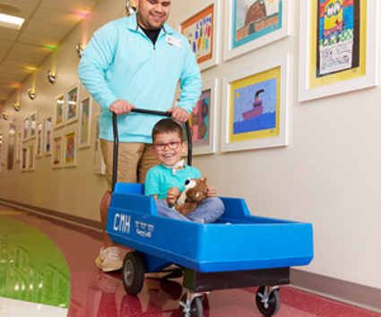 Child riding in a blue Children's Mercy wagon while being pushed down a hospital hallway by an adult