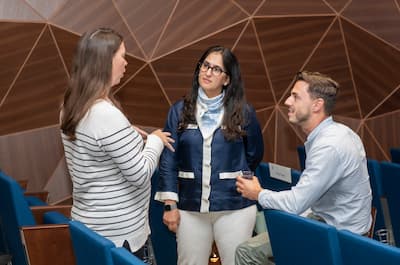 Three team members converse in a meeting room with blue auditorium seating.