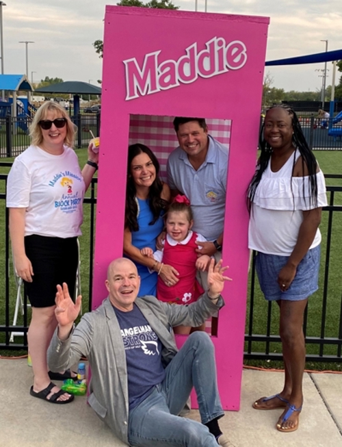 A man, woman, and female child stand in a lifesize Barbie box that says Maddie on the outside. Two women stand on either side of the box. A man sits in front of it with his hands in the air.