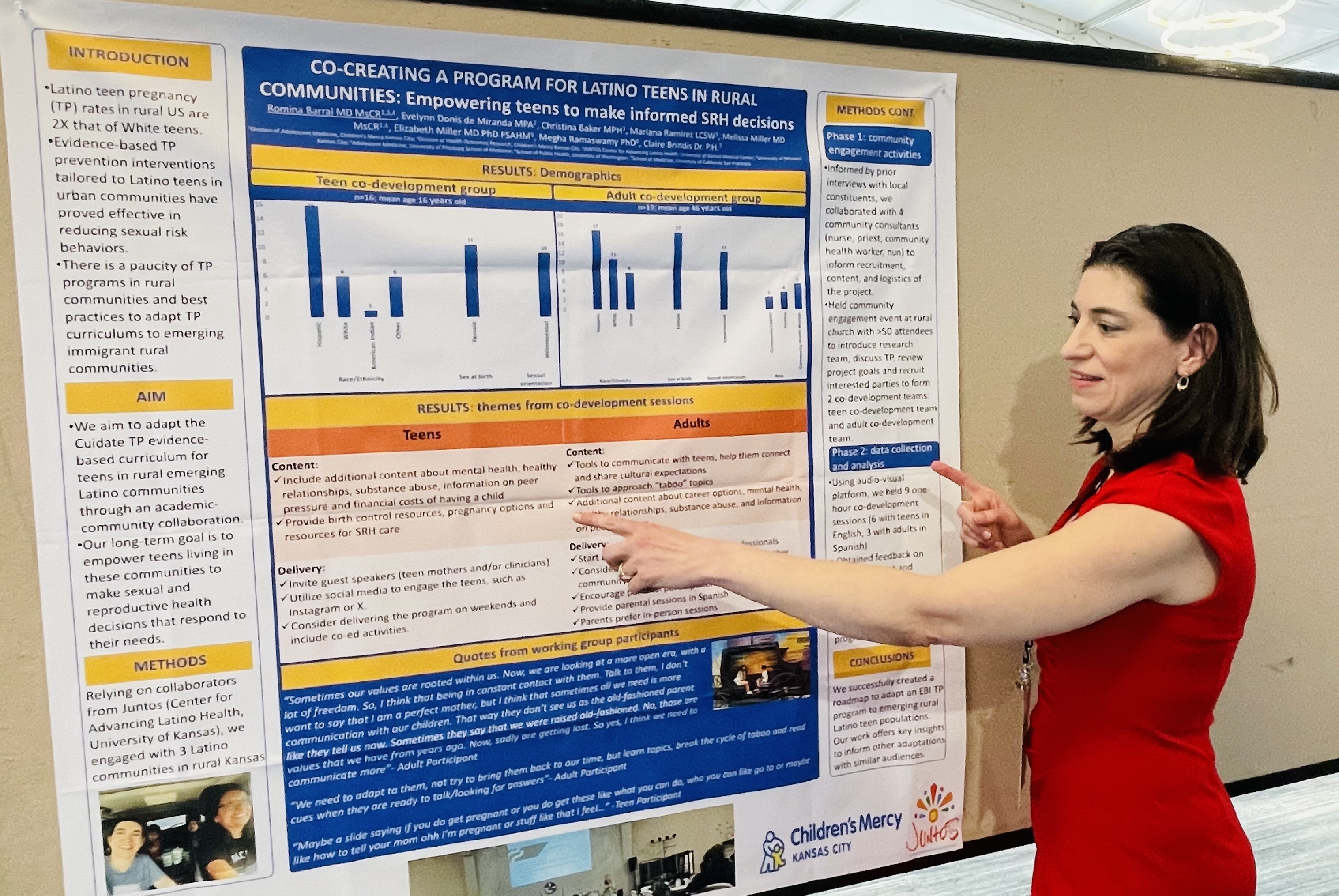 A woman wearing a red dress stands next to a research poster and points at something on it. The poster is titled Co-Creating a Program for Latino Teens in Rural Communities: Empowering Teens to Make Informed SRH Decisions.