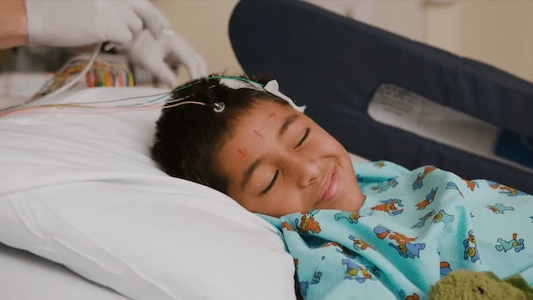 Child resting in a hospital bed while connected to monitoring electrodes on the scalp during a neurological evaluation