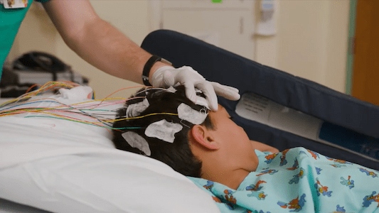 Child lying in a hospital bed with monitoring sensors attached to the head as a health care team member prepares diagnostic equipment