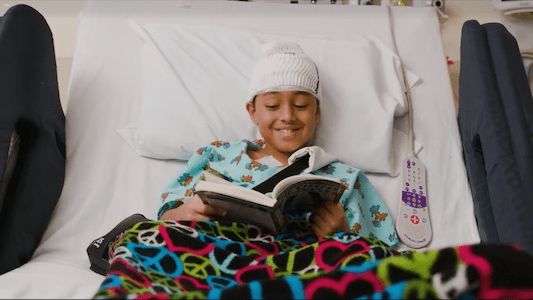 Child in a hospital bed wearing a neurological monitoring cap and reading a book