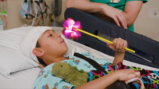 Child resting in a hospital bed while wearing a mesh cap that secures neurological monitoring sensors, holding a light-up toy wand