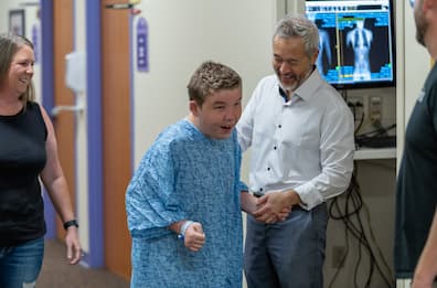 Patient wearing a hospital gown walking in a hospital hallway with support from a health care provider, while caregivers and team members look on during recovery and rehabilitation