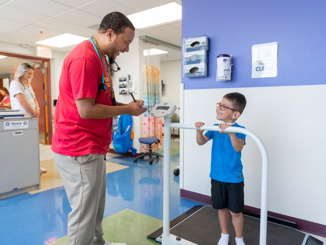 An adult health care worker talks with a young boy using a pediatric scale in a brightly lit clinic hallway