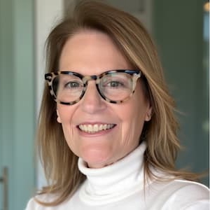 Professional headshot of a person wearing patterned eyeglasses and a white turtleneck sweater, photographed indoors against a softly blurred background.