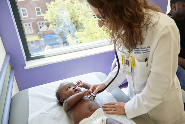 Health care provider using a stethoscope to examine an infant lying on an exam table during a pediatric clinic visit.