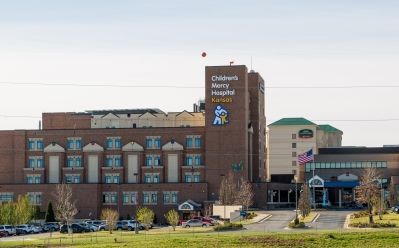Exterior view of Children’s Mercy Kansas hospital, showing the main entrance, multi-story brick building, drive-up entrance canopy, and hospital signage on a clear day.