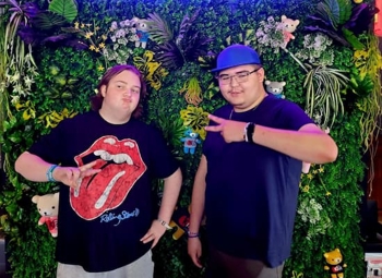 Two teenaged boys throw peace signs and goof off for the camera in front of a wall of hanging plants. 