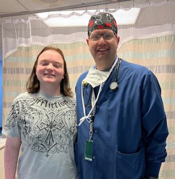 In a hospital room, a teenage boy and a doctor in blue scrubs pose and smile for a picture. 