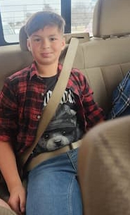 A 12-year-old boy is buckled into the back seat of a vehicle with a tan interior. He is smiling and wearing a red plaid shirt over a black tee shirt. 