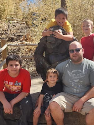 3 boys and their dad sit around a bronze sculpture of a gorilla at the zoo. A 4th boy has climbed up on the gorilla and is playfully covering its eyes. 