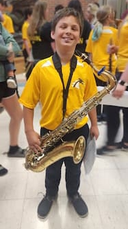A smiling 12-year-old boy wearing a yellow band t-shirt and holding a saxophone on a carrying strap around his neck. 