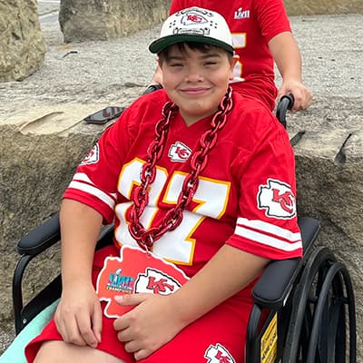 patient in wheelchair wearing  red Chiefs shirt, shorts, hat, and chain.  