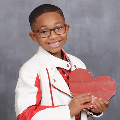 A boy in glasses poses in front of a photographer’s background. He's holding a wooden heart painted red and wearing a red and black checked shirt and white and red leather jacket.