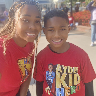A mom and son smile at the camera wearing coordinated red t-shirts. We can see an amusement park street behind them. The boy’s t-shirt has a cartoon version of himself and lettering that reads “Ayden: Kid Hero.”