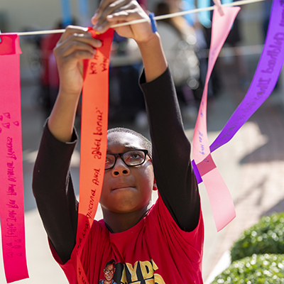 A boy in glasses concentrates as he attaches an orange ribbon with writing 	on it to a string. There are more people and ribbons out-of-focus behind him. He is wearing a t-shirt with a cartoon version of himself and lettering that reads “Ayden: 	Kid Hero.”
