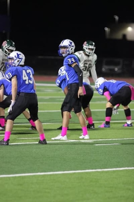 Youth football players lined up on the field during a nighttime game, wearing blue jerseys and pink socks.