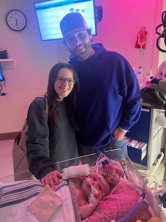Two adults standing beside an infant receiving care in a hospital bassinet, with medical monitoring equipment visible.