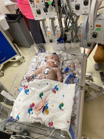 Infant lying in a hospital bassinet while connected to respiratory support, monitoring leads and other medical equipment.