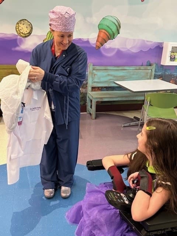 Health care team member in surgical scrubs showing a white coat to a child seated in a wheelchair in a colorful pediatric care area.