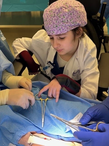 Young person wearing a surgical cap and white coat participating in a simulated medical procedure with guidance from gloved team members.