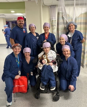 Child seated in a wheelchair surrounded by a group of hospital team members wearing surgical attire and caps in a pediatric hospital unit