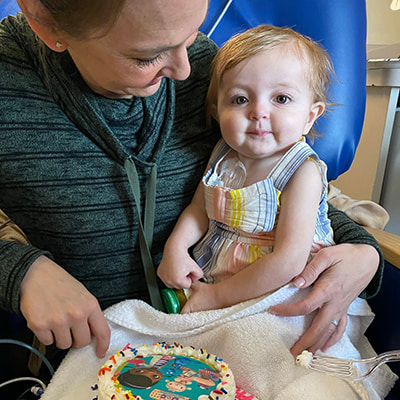 Infant sitting on an adult’s lap beside a decorated cake, with medical tubing visible.