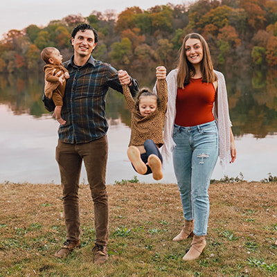 Two adults with two young children standing beside a lake, with autumn trees in the background and one child being lifted by the hands.