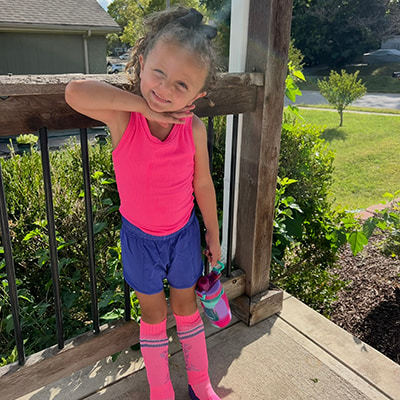 Child standing on a porch outdoors in athletic clothing while holding a water bottle