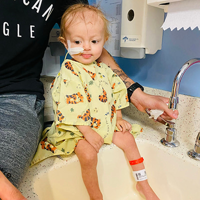 Young child wearing a hospital gown and nasal feeding tube sitting on the edge of a sink while an adult holds the child’s hand under running water.