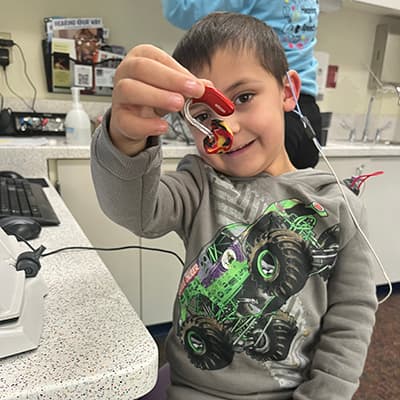 A young boy with brown hair shows his red hearing aid to the camera. He is in a clinic room wearing a gray sweat suit with a bright green monster truck on it.