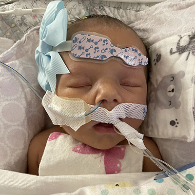 Close up of a small infant with medical tubes coming out of her nose and mouth. She has a medical monitor on her forehead beside a blue bow. She’s covered in soft blankets.  