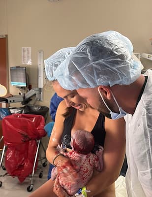 A person holds a newborn baby while another person in medical attire leans close beside them in a hospital room.