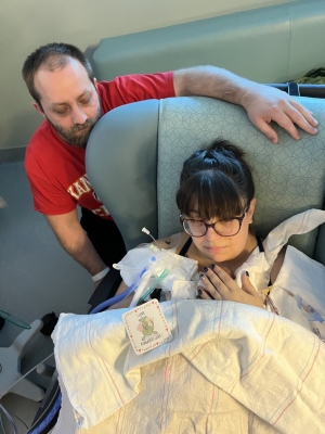 A mother with brown hair and glasses sits in a hospital chair with a barely visible baby on her chest. Many medical tubes and wires are coming out from under the blanket. A man in a red shirt looks on from behind the chair.