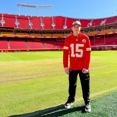 Young person wearing a red Kansas City Chiefs number 15 jersey standing on the field inside an empty football stadium.