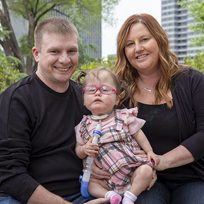 Two adults and a young child sitting together outdoors, with the child wearing pink glasses and using a tracheostomy tube.