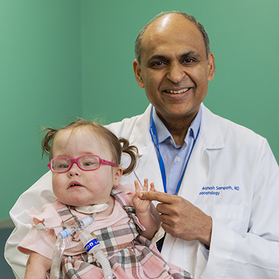 Young child wearing pink glasses and a tracheostomy tube posing with a health care provider in a white coat.